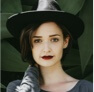 a woman in a black hat, infront of a leafy green background